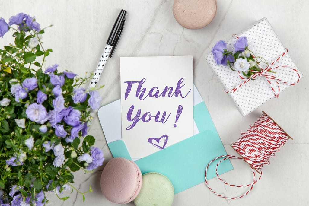 Thank you card with purple flowers and green leaves. 3 pastel macaroons sit on the table with a pen and a small box tied with red & white string. I'm thankful you are here.